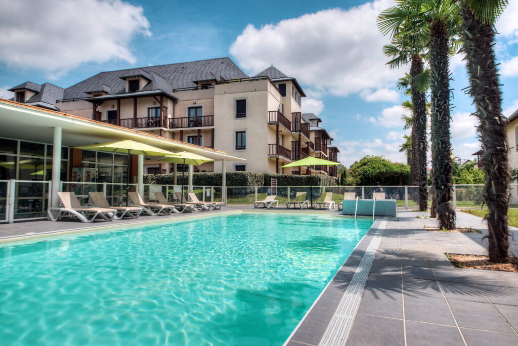 Piscine extérieure bordée de transats et de palmiers au Comfort Aparthotel Lourdes l'Acacia.