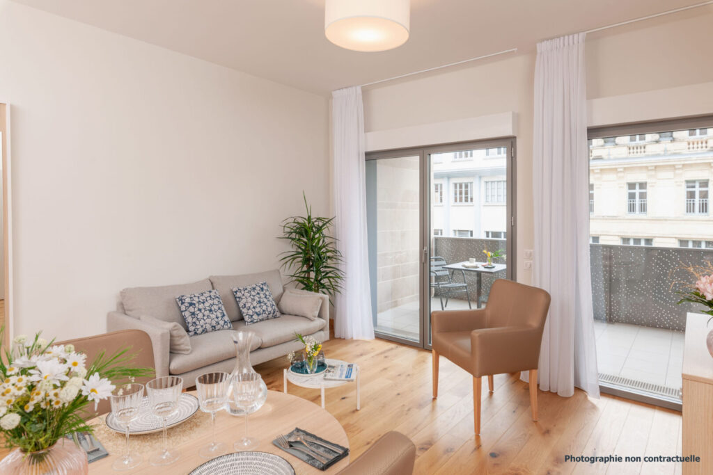 Salon lumineux avec canapé gris, table en bois et accès à un balcon.