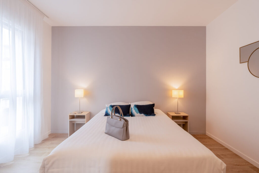 Chambre double spacieuse avec mur taupe, deux lampes de chevet et coussins colorés.