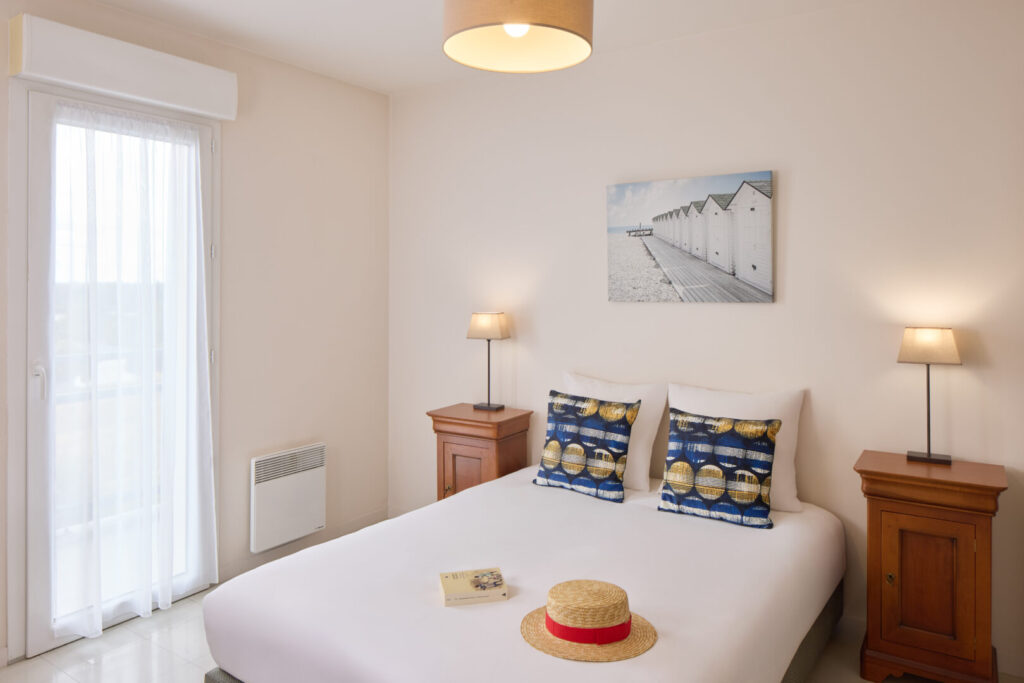 Chambre double lumineuse avec décoration marine, chapeau posé sur le lit et tableau mural.