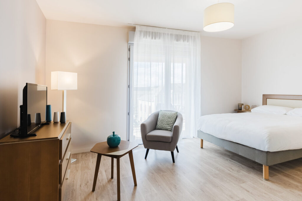 Chambre lumineuse, fauteuil design, table d’appoint et lit double près de la fenêtre.